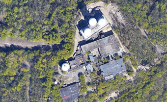 An aerial image of an artificial hill at Teufelsberg, Berlin. An ex-listening station, the building structure is now a gallery with a QR code painted on top, surrounded by greenery/forest.