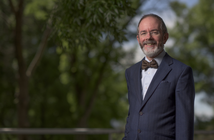 A/Prof. Douglas Guilfoyle is standing outside in front of greenery, wearing a suit and bow tie. He is smiling.
