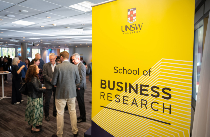 Image of UNSW Canberra branded banner with people networking behind the banner.